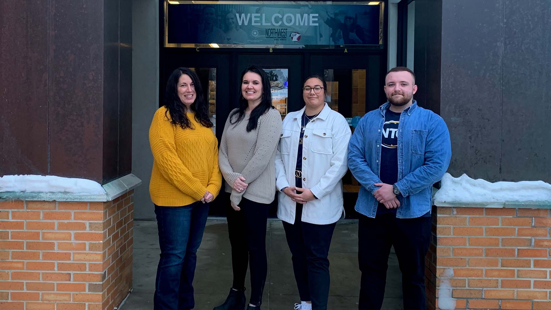 Four people standing in front of Northwest Technical College: a woman in a yellow sweater; a woman in a beige sweater; a woman wearing an NTC t-shirt with a white jacket over it; and a man with a blue NTC t-shirt with a blue denim shirt over it.