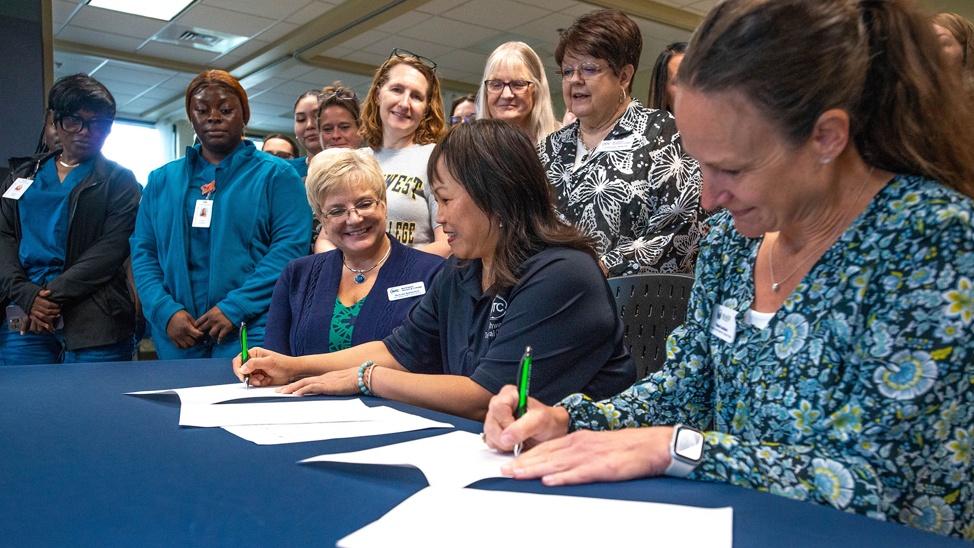 Three BSU and NTC administrators sit at table and sign contracts. Many NTC nursing students are watching from behind them.