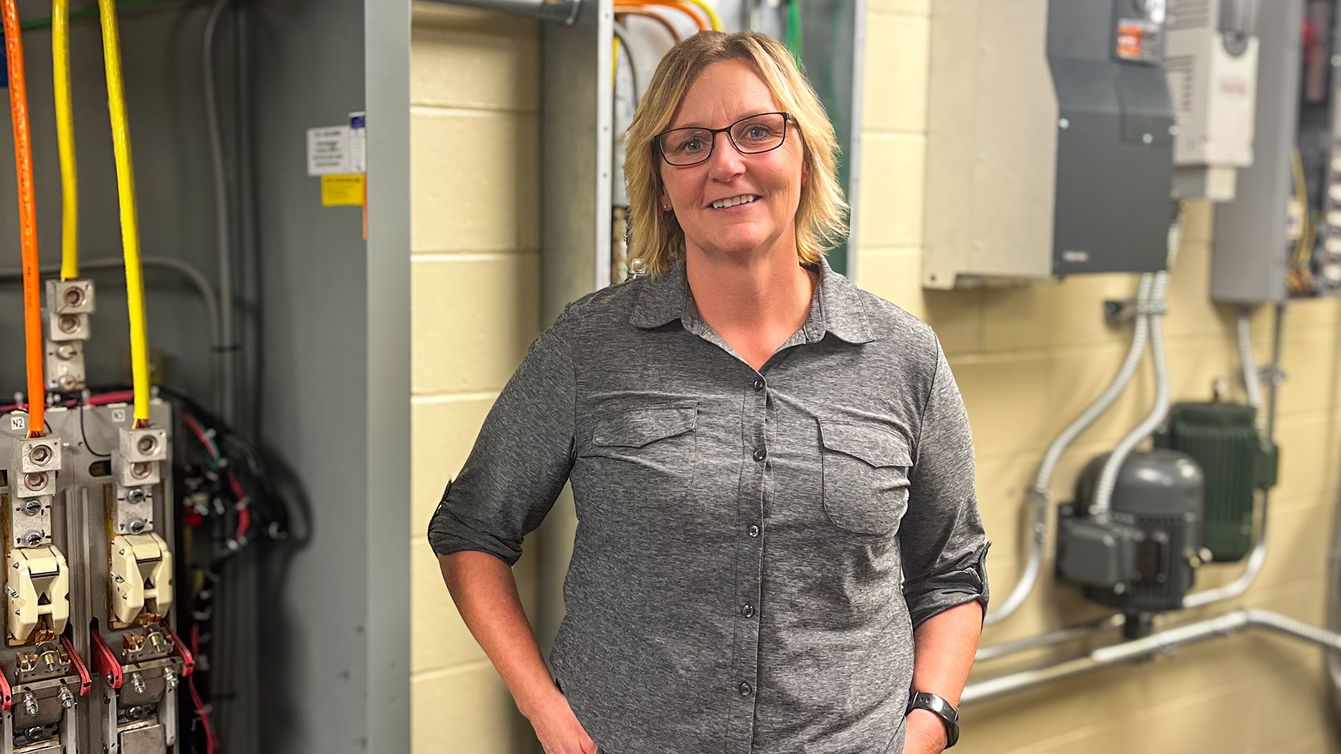 A woman with blonde hair wearing glasses and a grey long-sleeved shirt with the sleeves roleld up stands in front of several electrical conduit boxes