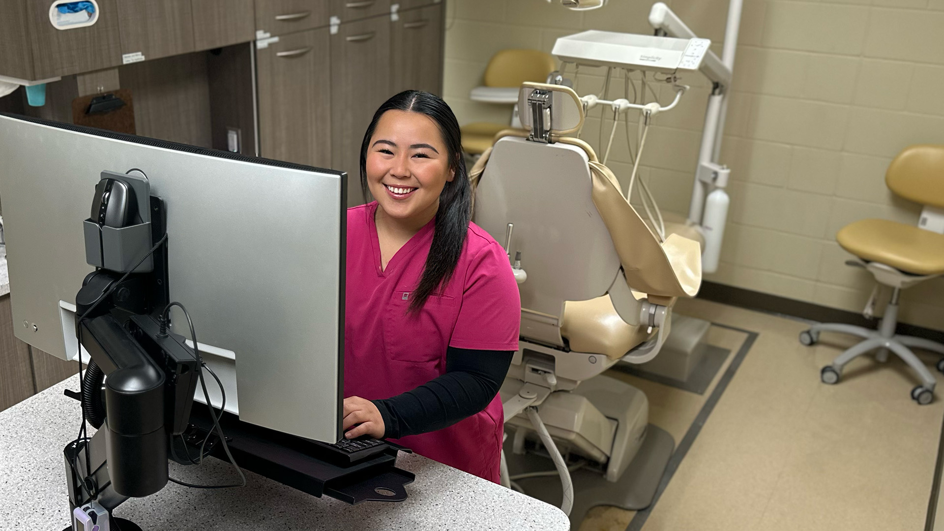 A student in fuscia scrubs over a long-sleeved black shirt smiles from behind a computer monitor. A dental exam chair is behind her.
