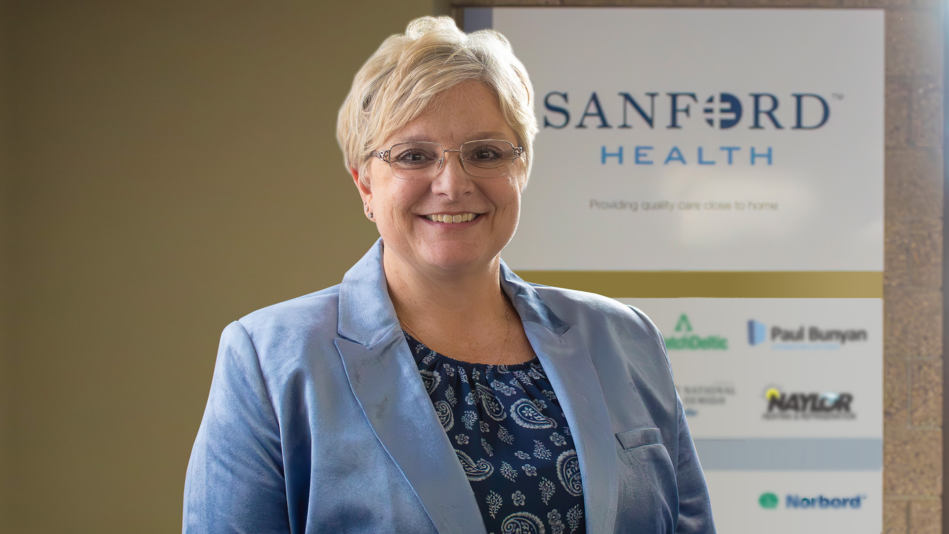 A woman with short blonde hair and glasses is wearing a shiny, light-blue blazer over a patterned dark blue shirt. The wall behind her has a large "Sanford Health" logo.