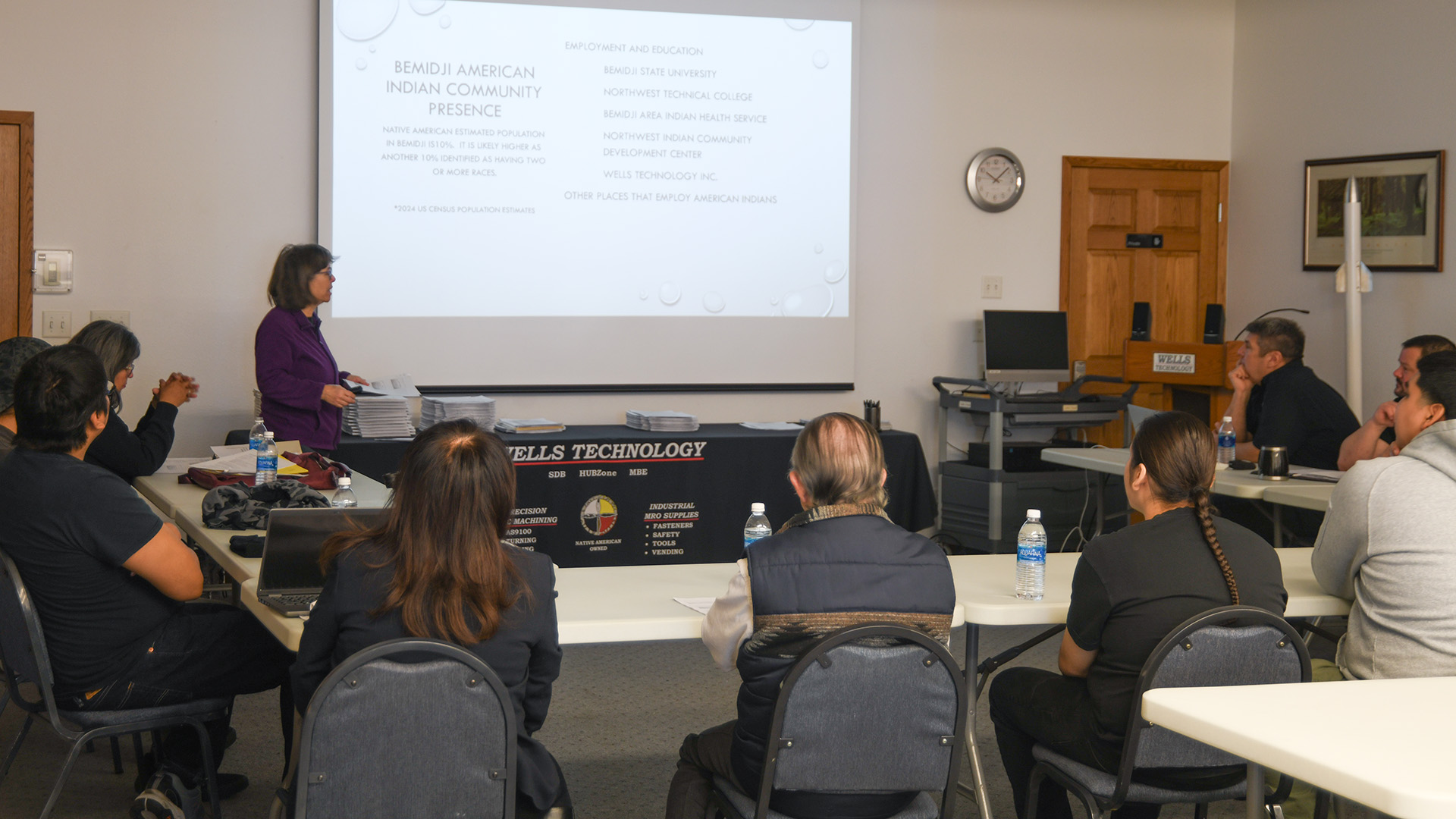 Dr. Pauline Danforth speaks to Wells Academy students in a conference room. A PowerPoint is running on a large screen behind her. She's wearing a purple jacket.