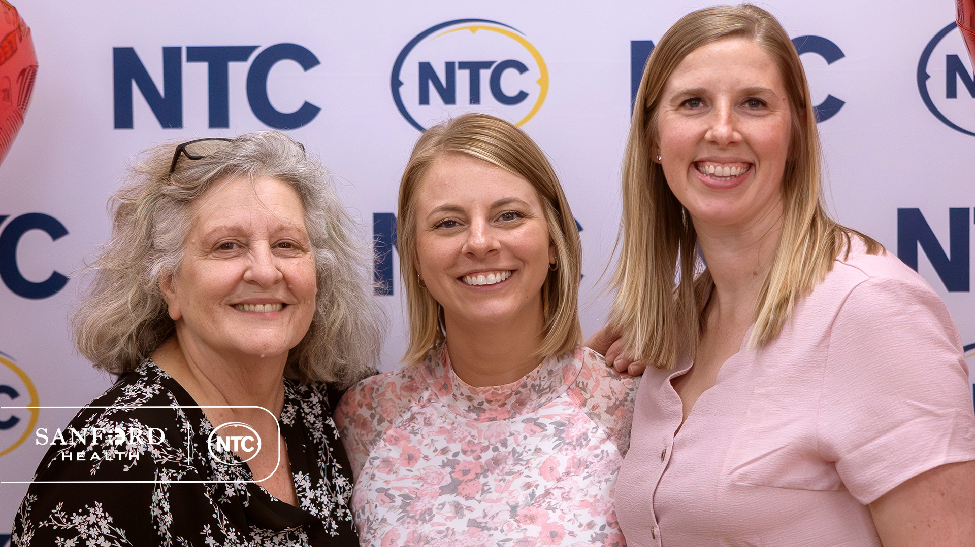 Mindy Williams and Maggie Wolverton stand next to Kaileigh Rybak in front of a backdrop covered in NTC logos.