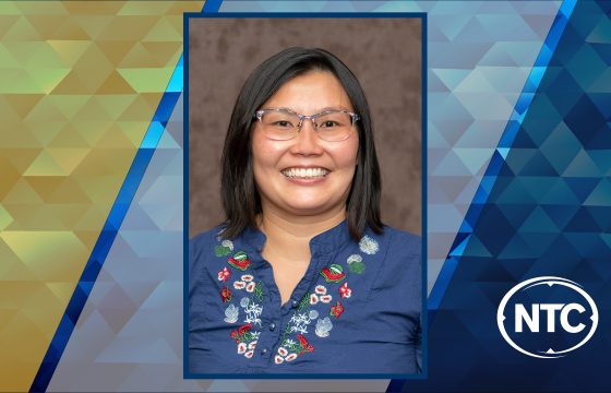 photo of a smiling woman with black hair wearing glasses and a blue embroidered shirt with NTC colors in the background