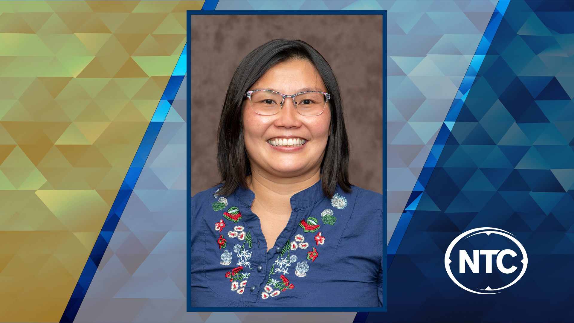 photo of a smiling woman with black hair wearing glasses and a blue embroidered shirt with NTC colors in the background