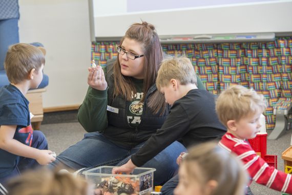 An NTC student holding up a gluestick in a classroom of young children playing