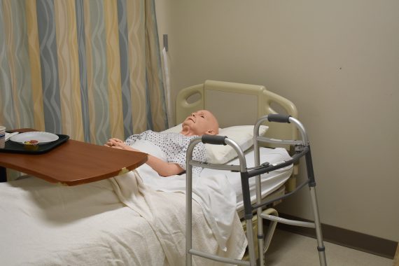 A mannequin of an elderly man a bald head is tucked into a hospital bed in the Sanford/NTC Health Sciences Lab for CNA students at Bemidji High School. A walker sits next to the bed.