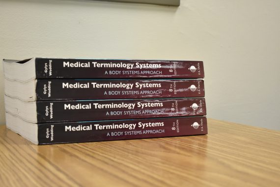 A stack of four maroon textbooks with "Medical Terminology Systems" written on the spines in white letters