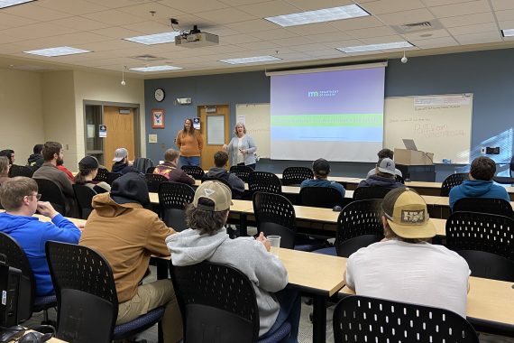 Two people stand in front of a classroom with a PowerPoint presentation on a large screen. Many NTC students are watching their presentation.