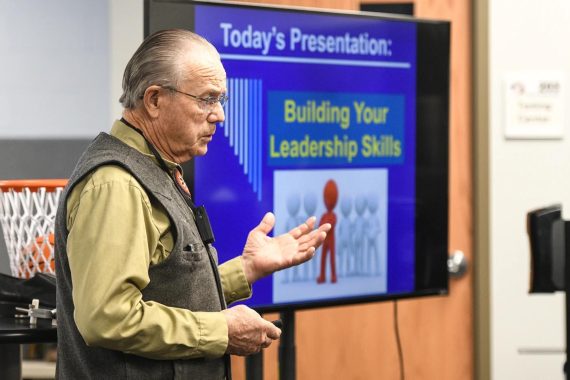 A man with grey hair and glasses, wearing a beige long-sleeved shirt and dark vest, speaks in front of a PowerPoint presentation.