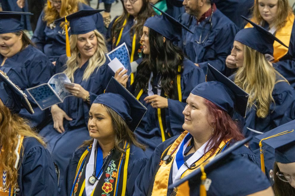 Students in blue caps and gowns, several with yellow cords and stoles, are graduating from Northwest Technical College