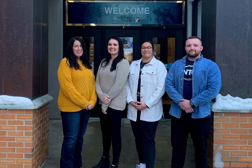 Four people standing in front of Northwest Technical College: a woman in a yellow sweater; a woman in a beige sweater; a woman wearing an NTC t-shirt with a white jacket over it; and a man with a blue NTC t-shirt with a blue denim shirt over it.
