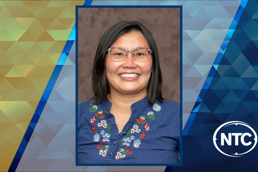 photo of a smiling woman with black hair wearing glasses and a blue embroidered shirt with NTC colors in the background