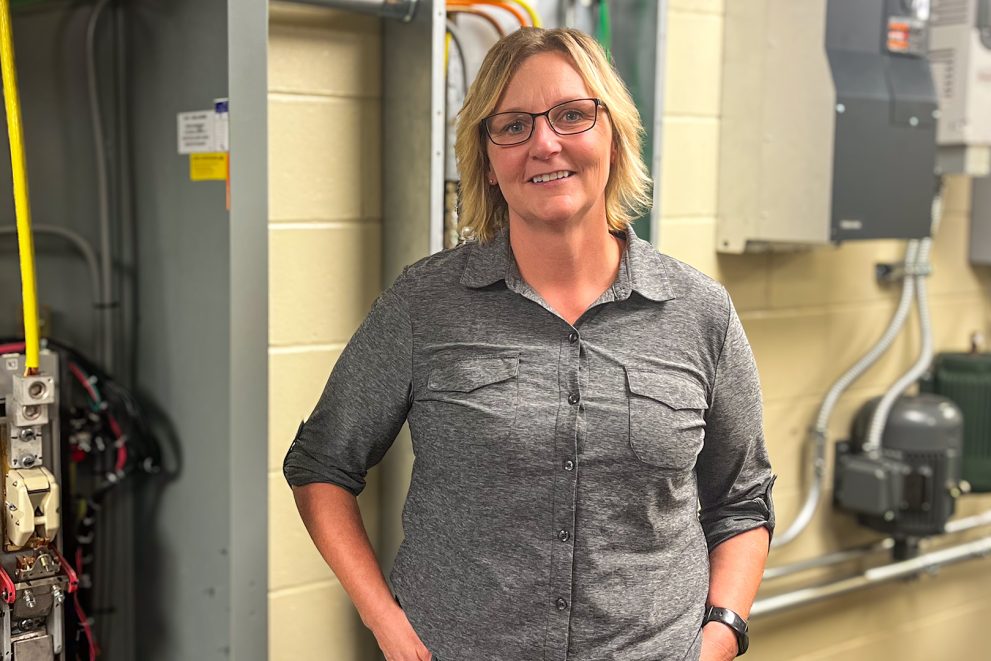 A woman with blonde hair wearing glasses and a grey long-sleeved shirt with the sleeves roleld up stands in front of several electrical conduit boxes