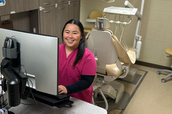 A student in fuscia scrubs over a long-sleeved black shirt smiles from behind a computer monitor. A dental exam chair is behind her.
