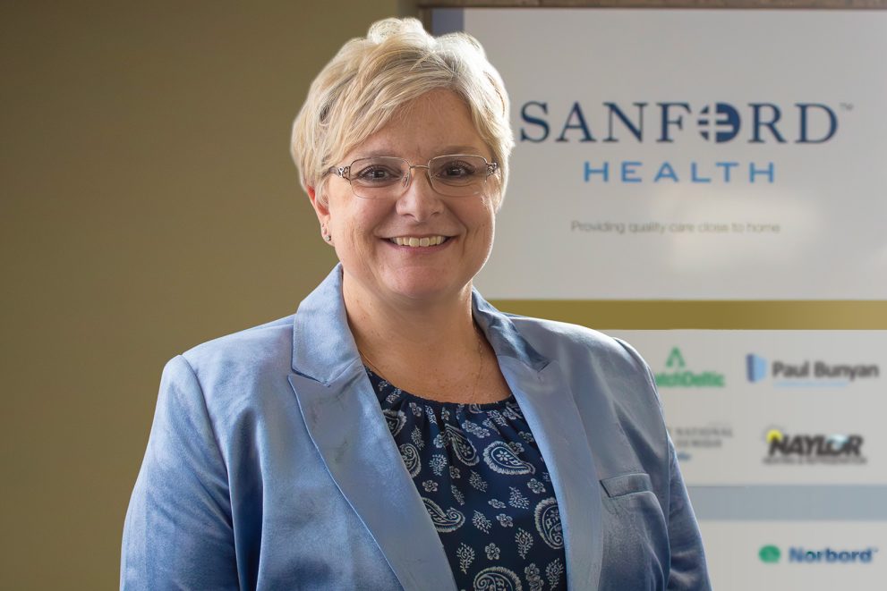 A woman with short blonde hair and glasses is wearing a shiny, light-blue blazer over a patterned dark blue shirt. The wall behind her has a large "Sanford Health" logo.