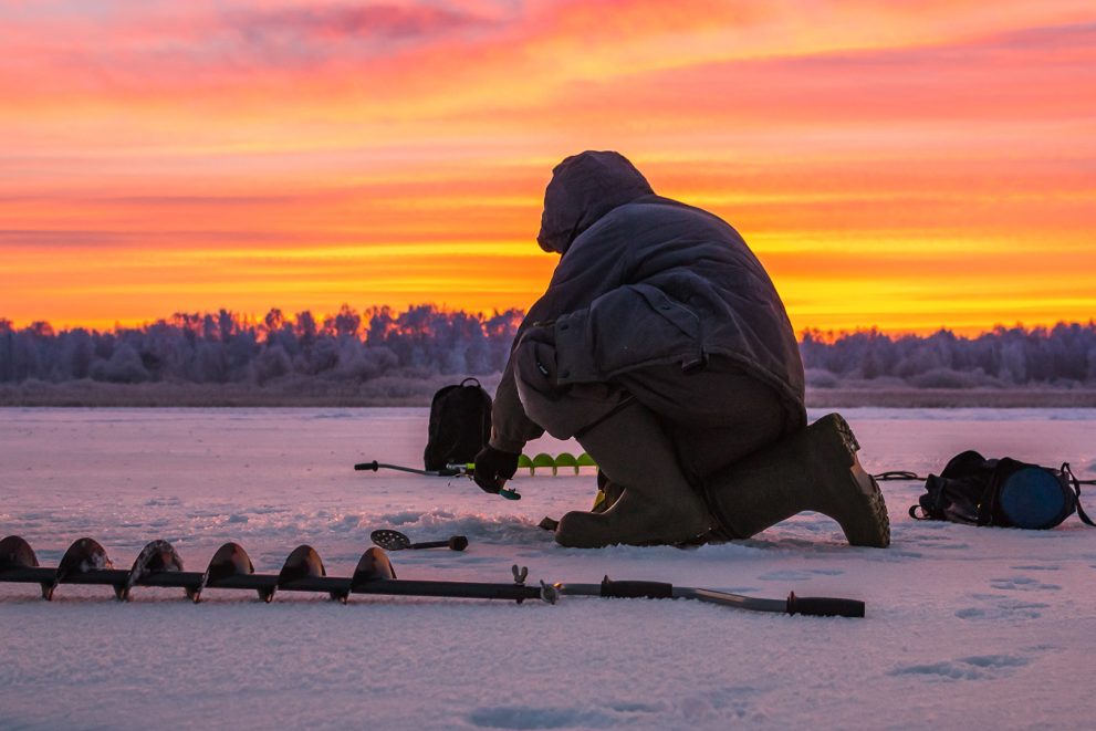 A stock photo of an ice fisherman with an auger. The sky in the photo is brilliant orange.