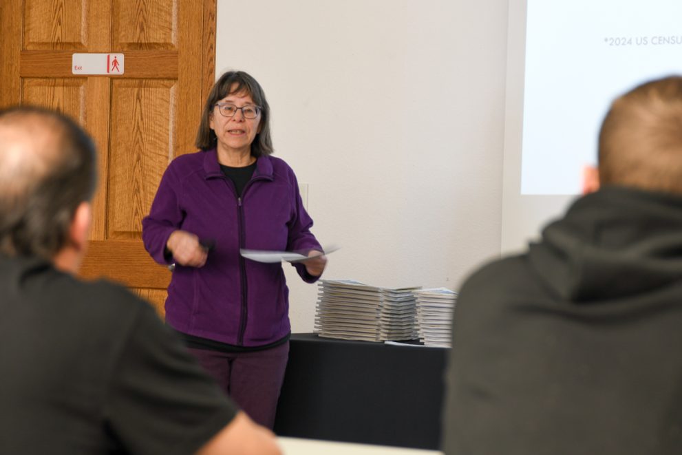 Dr. Pauline Danforth speaks to Wells Academy students in a conference room. A PowerPoint is running on a large screen behind her. She's wearing a purple jacket.