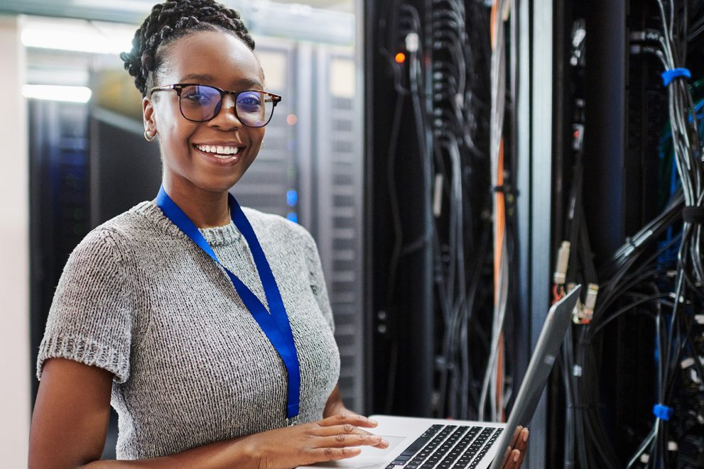 A stock photo of a woman wearing glasses and holding a laptop computer in her hands. She is in a server room.